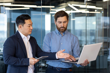Two young interracial businessmen standing in office, working on laptop, talking and discussing...
