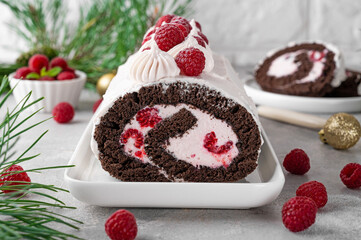 Chocolate christmas roll cake with cream and fresh raspberries on a white plate on a gray...