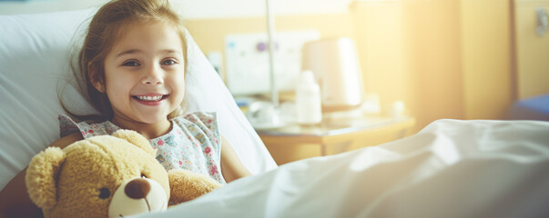 Small young girl smiling in hospital room with brown teddy bear on her lap. - obrazy, fototapety, plakaty