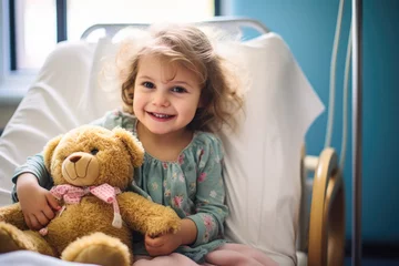 Fotobehang Small young girl smiling in hospital room with brown teddy bear on her lap. © PixelGallery
