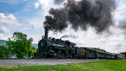 A View of a Narrow Gauge Restored Steam Passenger Train Blowing Smoke, Starting To Pull Out of a Station on a Summer Day