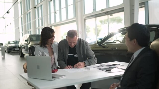 Middle aged couple signing contract on new car and getting keys from salesmen