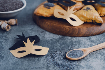 Jewish carnival Purim celebration on hamantaschen or haman's ears cookies decorated with DIY carnival masks on a wooden board. Festive sweet or salty pastries with poppy seeds.