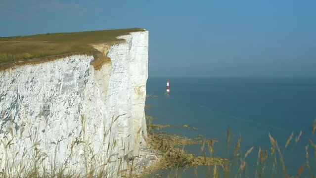Lighthouse at the end of white walls topped with grass rising above blue sea. Crumbling chalk sea cliffs stretching along picturesque coastline in southern England on a beautiful sunny summer day.