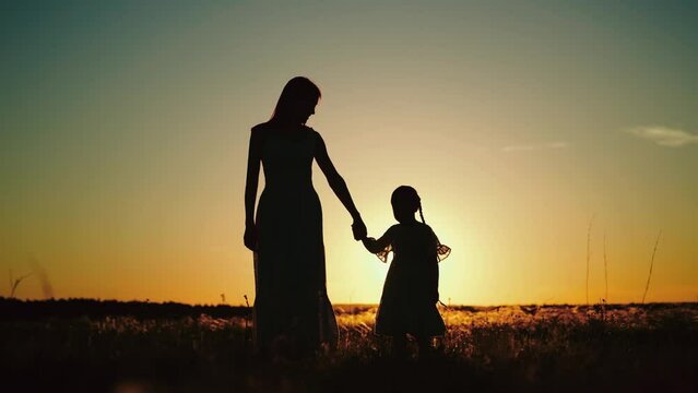 Silhouettes of mamma holding hand of daughter and talking about childhood while walking with child in sunlit field. Silhouettes of mamma with daughter sharing secrets at setting sun in park