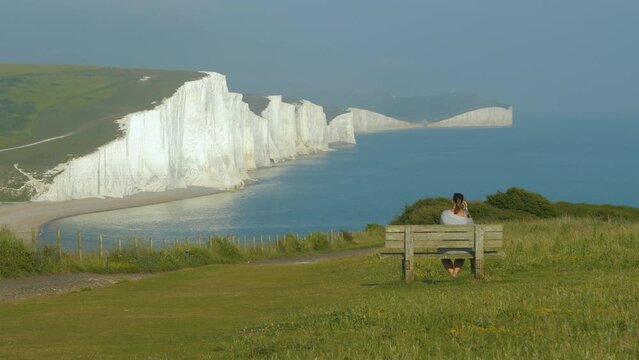 Traveller sitting on wooden bench and taking photos of beautiful white cliffs. Young woman is travelling and exploring picturesque coastline in Southern England. Vantage point with magnificent view.