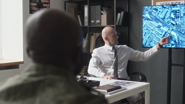 Over shoulder of military professor in formalwear pointing at satellite map on screen and asking unrecognizable cadet to answer during seminar in service academy