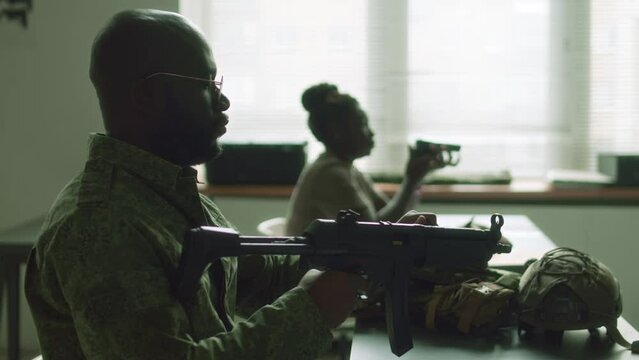 Side chest up of African American male cadet in glasses and camouflage uniform learning how to use machine pistol during class in military college
