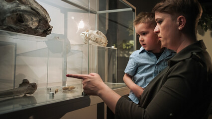 Curious little boy in his mothers arms looking at an animal skeleton in a bone display case at a natural history museum exhibition.