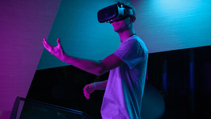 Young man with a VR headset having an immersive virtual reality experience in a room with purple lighting, making various hand movements in the air.