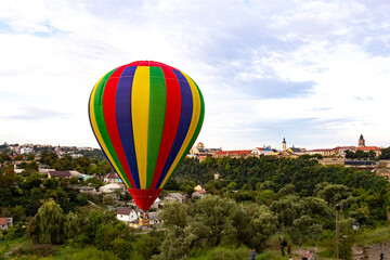 Multi colored hot air balloon flying over the city with a lot of green trees. Balloon basket with people. Romantic trip. Copy space