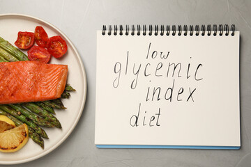 Notebook with words Low Glycemic Index Diet and plate of tasty grilled salmon on grey table, flat...