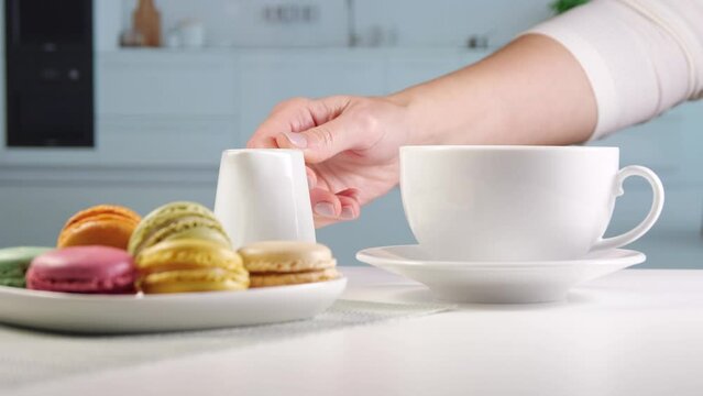 Woman pours milk from a milk jug into a coffee cup. Breakfast table in bright kitchen. Coffee with milk and colorful macarons. Sugar consumption and coffee consumption