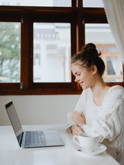 Creative young woman working at a desk at home in a cozy interior with a cup of drink, home office freelancer with a smile, communication and education online