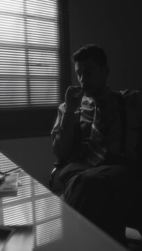 Vertical monochrome pan shot of pessimistic middle-aged man in officewear smoking and then crushing cigarette in ashtray sitting near window in room