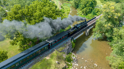 An Aerial View of a Narrow Gauge Steam Passenger Train, Traveling Crossing a Bridge Over a Stream, Blowing Smoke, on a Sunny Summer Day