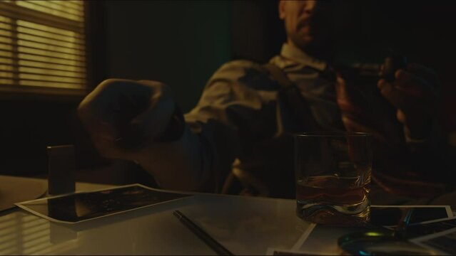 Medium shot of unrecognizable bearded man sitting alone at table with glass of liquor lighting up and smoking pipe and feeling nostalgic while looking at picture in dark room