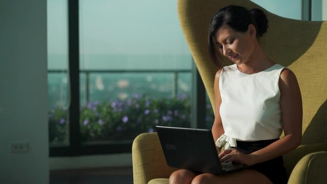 Young Asian business woman wear suit sit on couch chair working on laptop in modern modern business lounge office. Gorgeous lady freelancer digital nomad concept. Working online. Urban background