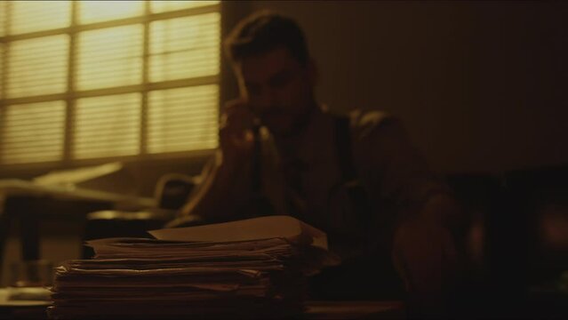 Sepia closeup of pile of letters lying on table with bearded man speaking on mobile phone sitting on sofa in background in dimly lit room