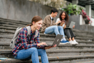 Happy diverse university students sitting on steps, using laptops and tablets, enjoying conversation. Student exchange and study abroad program, Asian man, african american woman and caucasian people. - obrazy, fototapety, plakaty