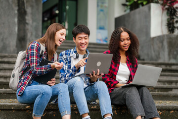 Happy diverse university students sitting on steps, using laptops and tablets, enjoying conversation. Student exchange and study abroad program, Asian man, african american woman and caucasian people.