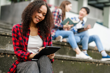 Happy diverse university students sitting on steps, using laptops and tablets, enjoying...