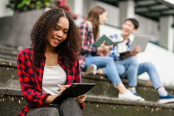 Happy diverse university students sitting on steps, using laptops and tablets, enjoying...