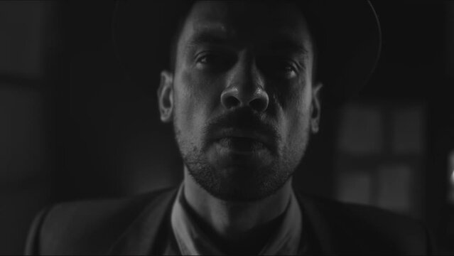 Monochrome portrait of serious Caucasian bearded bandit-like man in hat lifting up head and staring at camera indoors