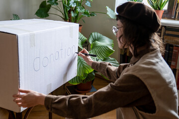 Girl volunteer preparing box for donations writing inscription. Altruist student going to hold...