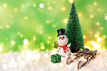 Cheerful snowman with a wooden sleigh and a Christmas tree on artificial snow and a green background with bokeh. New Year, Christmas, Christmas Eve.