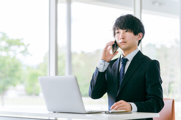 スマートフォンで話しながらパソコンを使う若いアジア人ビジネスマン
 - obrazy, fototapety, plakaty