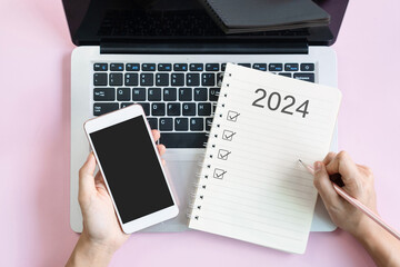 Flat lay of woman hand writing 2024 to do list on notebook while holding mobile phone with blank screen. Copy space, top view. 2024 planning concept