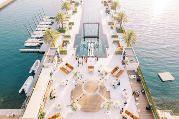 Festive tables stand on a wide pier with a swimming pool and moored yachts. Drone