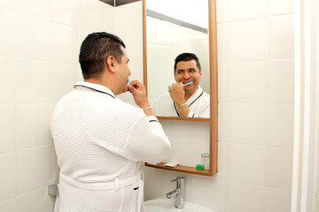 Dark-skinned 40-year-old Latin man brushes his teeth in front of the bathroom mirror in a bathrobe...