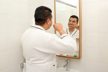 Dark-skinned 40-year-old Latin man brushes his teeth in front of the bathroom mirror in a bathrobe...