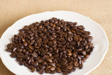 Fototapeta premium coffee beans. on a white large plate lies a large number of small brown coffee beans, the plate stands on the table, top view