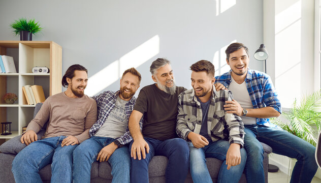 Portrait of a male bearded friends sitting on sofa, smiling and having fun at home in the living room. Photo of group of brutal friendly men in casual clothes hugging indoors on the party.
