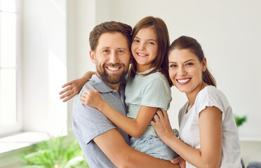 Close up portrait of a young happy family of three standing at home with their child girl looking at camera and smiling. Joyful parents hugging their daughter. Love, care and happy family concept.