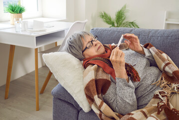 Sick senior woman measures her temperature. Mature lady has seasonal cold, flu and fever. Older adult patient in warm blanket and scarf lying on sofa at home and looking at thermometer in her hands - Powered by Adobe