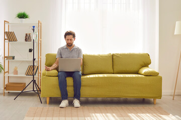 Bearded young man sitting on the couch at home and working with laptop in the living room while...