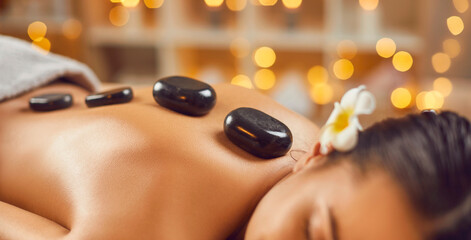 Close up photo of female back with hot stones. Portrait of pretty young brunette woman with closed eyes lying alone relaxing in spa salon getting massage therapy. Wellness and beauty day concept. - obrazy, fototapety, plakaty