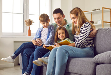 Smiling, happy family with two children are engaged in educational activities. Loving young parents with their children read book together, sitting at home on cozy sofa in the living room.