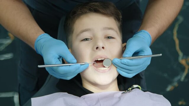 Male hands in latex gloves examine lower teeth of kid using tooth scraper, concave mirror, patient sitting calmly in dentist chair with napkin on chest. High quality 4k footage
