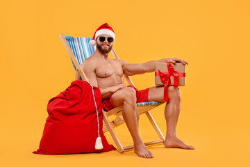 Muscular young man in Santa hat with deck chair, bag, sunglasses and Christmas gift box on orange...