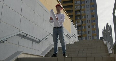 Attentive architect in helmet carefully reviews construction plan descending stairs of building. Expert control of adherence to drawing plan concept