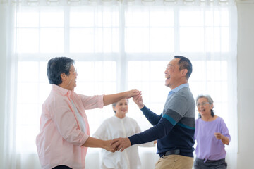 Obraz premium Asian Older male and females people dance with their partners on a dancing floor in living space. Happy older couple performing get exercise. Joyful carefree retired senior friends enjoying relaxation