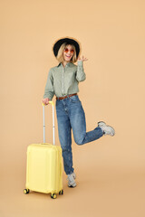 Happy pretty gen z blonde young woman, smiling cool girl tourist wearing hat and sunglasses with yellow travel suitcase waiting for vacation standing isolated on beige background. Full length.