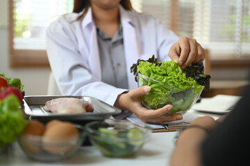 Professional nutritionist in white coat consulting client at table with organic vegetables and...