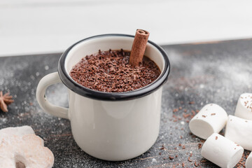 Homemade spicy hot chocolate drink with cinnamon stick, grated chocolate in enamel cup on wooden table with cookies, white marshmallows and star anise