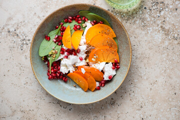Rustic plate with persimmon, goat cheese, spinach and pomegranate salad, above view on a beige granite background, horizontal shot
